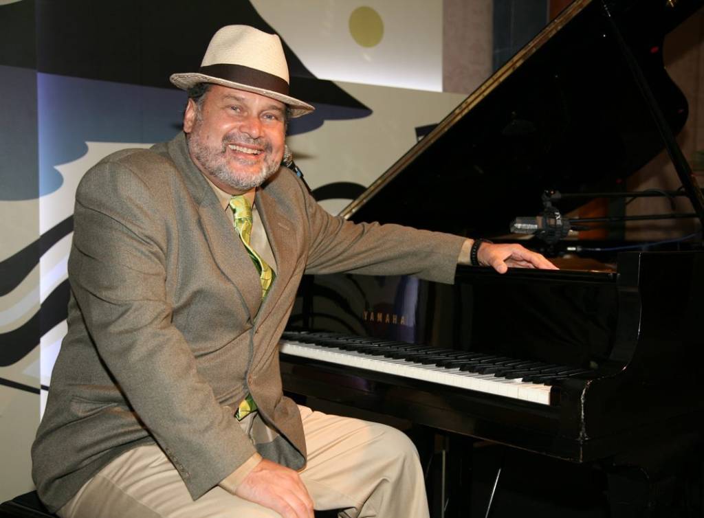 O pianista Marcos Ariel está de paletó cinza, gravata amarela, calça cinza-claro e chapéu panamá branco com faixa preta. Ele está sorrindo, sentado e com sua mão esquerda apoiada em um piano de cauda.
