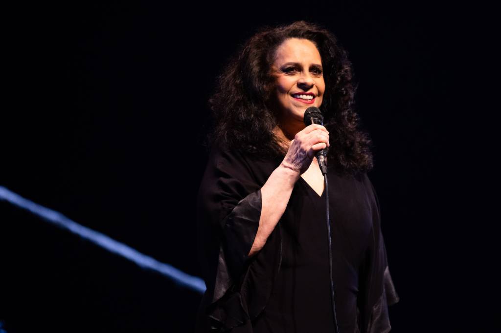 A cantora Gal Costa de vestido preto no palco com fundo preto. Ela segura o microfone e está sorrindo.