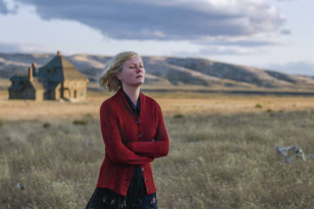 A atriz Kirsten Dunst em um campo gramado. Ela usava casaco vermelho, saia preta, está com os braços cruzados e os cabelos ao vento