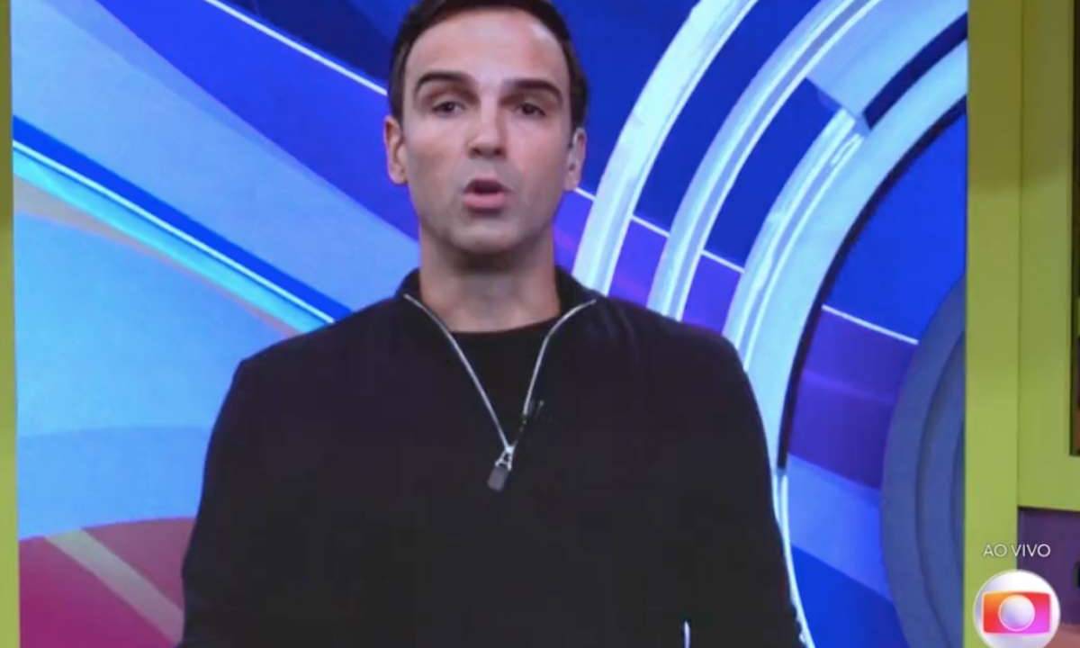 O apresentador Tadeu Schmidt de blusa preta de manga e com colar microfone do BBB. Ao fundo, a marca do programa, em tons de azul.