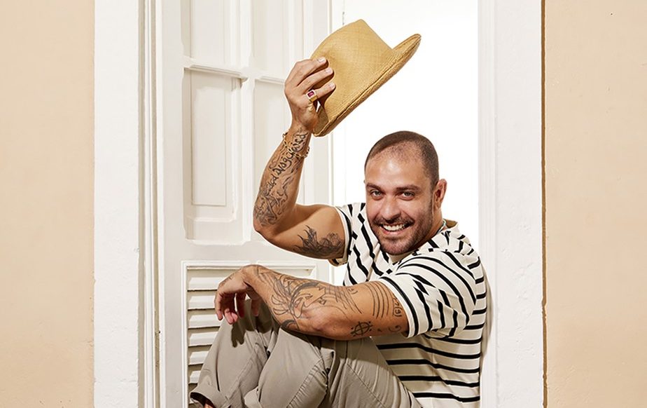 O cantor Diogo Nogueira está sentado no batente de uma janela de madeira branca. Ele veste calça bege e camisa branca com listras pretas e segura um chapéu na mão direita, acima da cabeça.