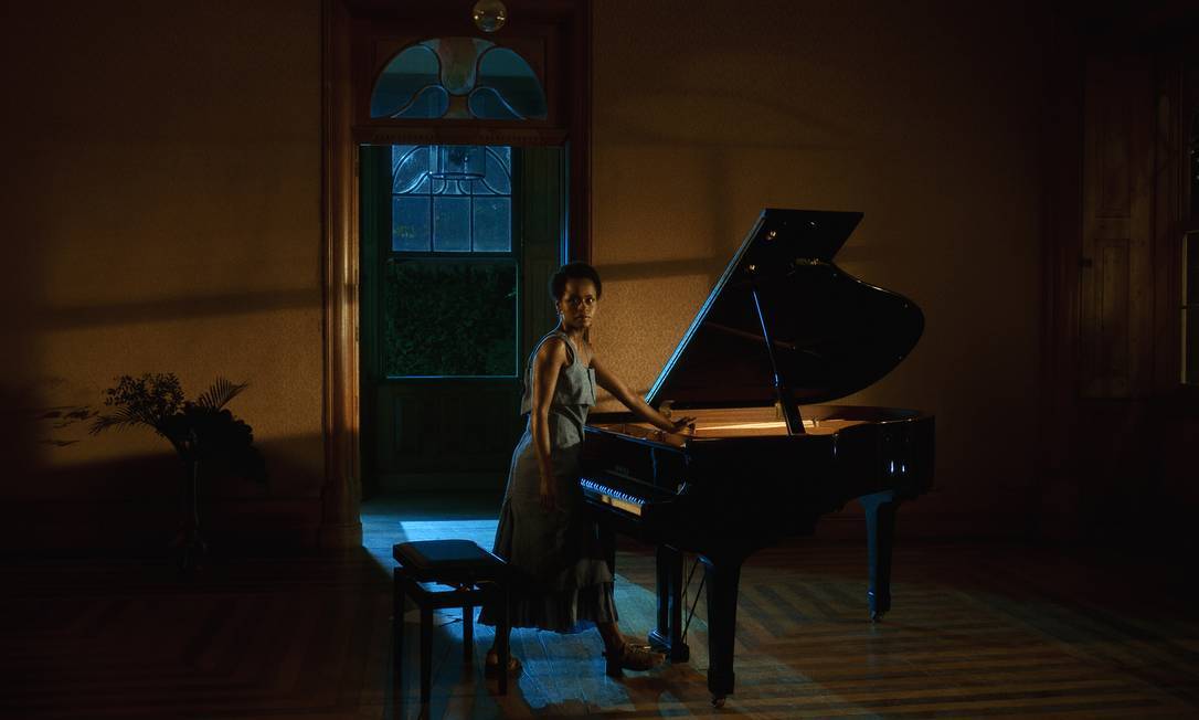 A atriz Raquel Paixão é negra, tem os cabelos curtos, usa vestido marrom e está de pé, diante de um piano de cauda. Ela está virada para a câmera. mas olha para seu lado direito.