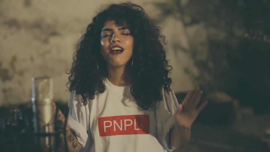 A cantora Marina está cantando. Ela está com os cabelos castanhos cacheados soltos, cantando, usa batom vermelho e blusa branca com uma tarja vermelho onde está escrito PNPL.
