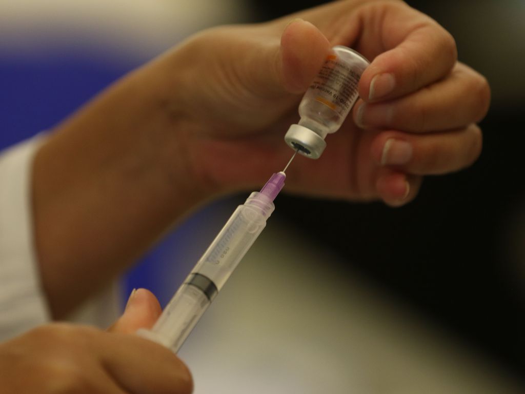 Uma das mãos de uma pessoa branca segura um frasco de vacina enquanto a outra mão segura a seringa, retirando a dose para ser aplicada