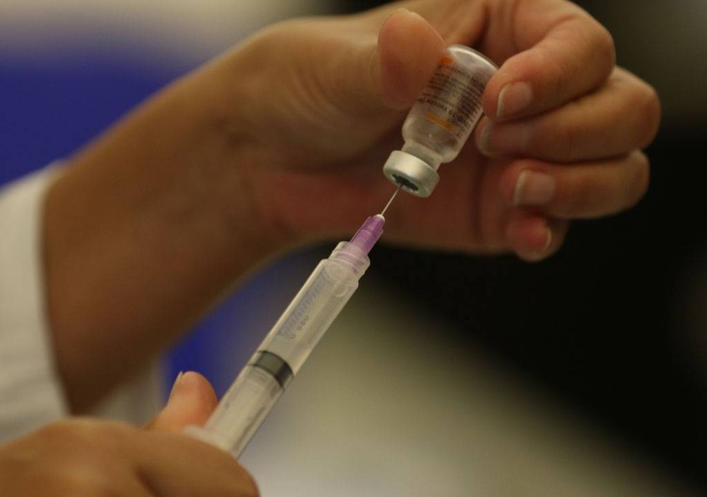 Uma das mãos de uma pessoa branca segura um frasco de vacina enquanto a outra mão segura a seringa, retirando a dose para ser aplicada
