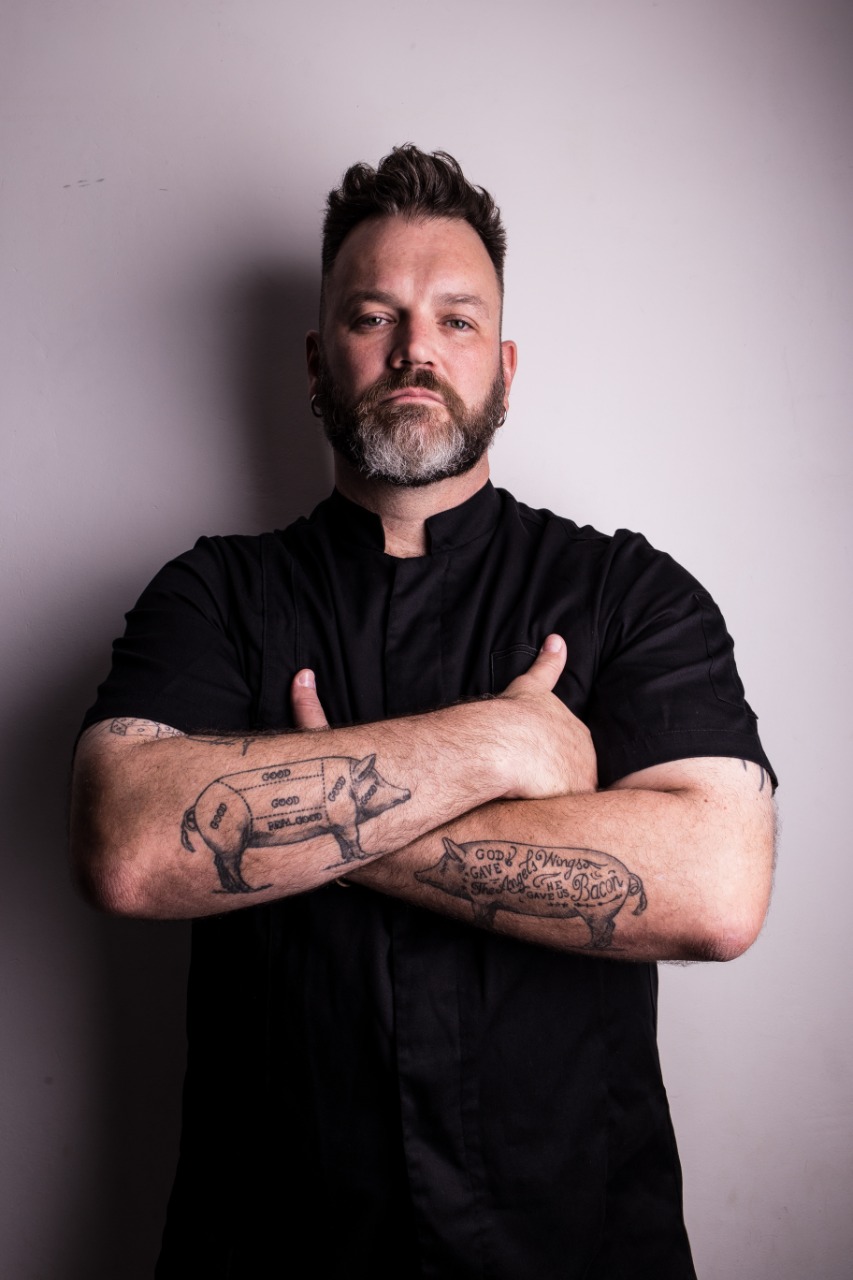 Foto mostra chefe de cozinha com barba e uniforme preto com os dois braços cruzados e com tatuagens de dois porcos