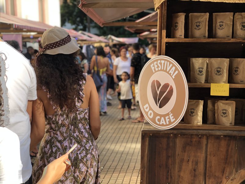 Foto mostra casal andando em frente a uma bacarra com grãos de café à venda e uma placa redonda escrita "Festival do café", com símbolo de um coração feito de dois grãos de café
