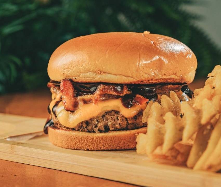 Foto de um hambúrguer em cima de uma tábua com batatas hashtag à direita, em primeiro plano