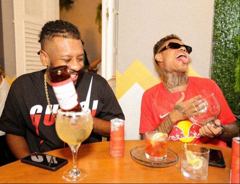 Os rappers BK' e MC Cabelinho estão sentados à mesa, sorrindo. Cada um tem um drinque à sua frente.