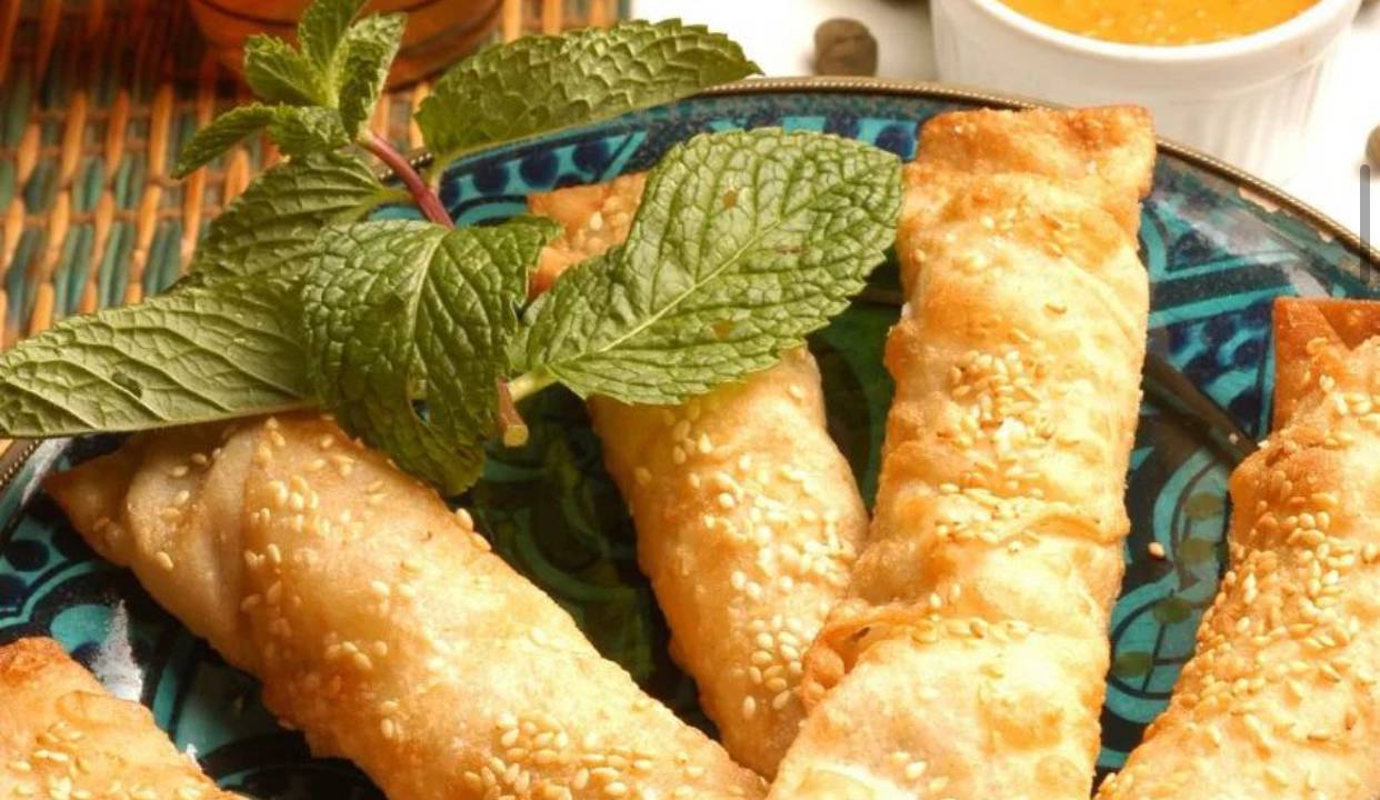 Foto mostra cinco mini pastéis compridinhos empilhados acompanhados de um molho na cor laranja