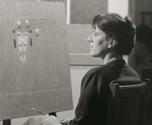 Imagem em preto e branco. A artista Eleonore Koch está de lado, olhando para uma tela em um cavalete à esquerda fa foto. Ela segura um pincel com a mão esquerda, tem os cabelos pretos, usa camisa de manga 3/4 escura e saia abaixo dos joelhos clara.