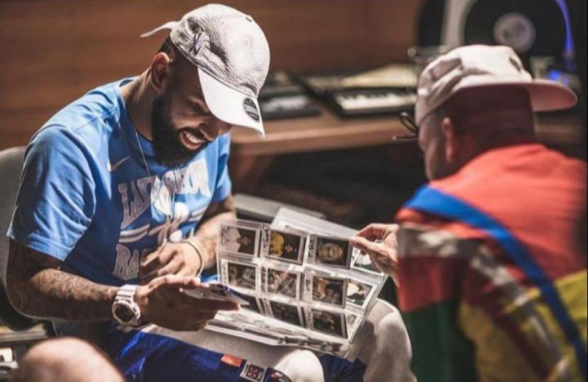 Gabigol está sentado, de casaco de moletom azul, calça de moletom azul e cinza, e boné bege, vendo um álbum, enquanto Papatinho está quase de costas para a foto, de frente para o jogador, usando moletom vermelho e azul e boné bege com a aba virada para trás.