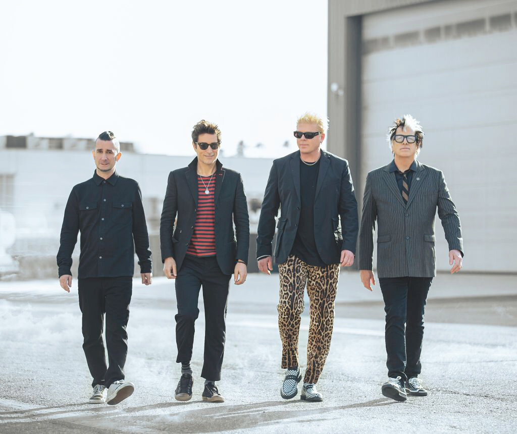 Os quatro integrantes do grupo The Offspring estão de pé, em uma área externa. Todos
