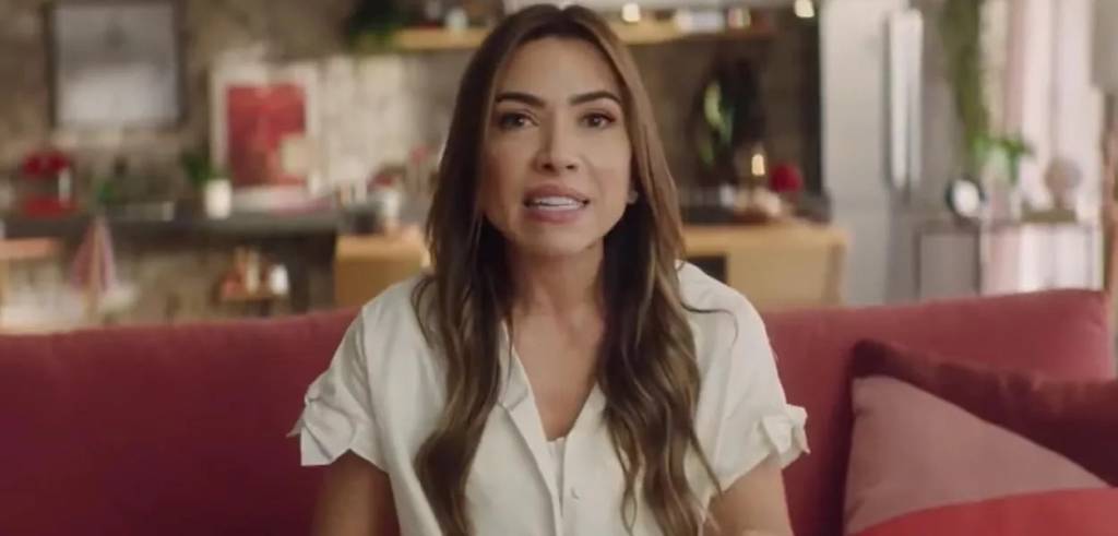 patricia-abravanel Patrícia Abravanel está falando. Ela usa camisa branca de manga curta sobre top branco. Ao fundo, um sofá vinho e objetos como escrivaninha, cadeiras e estantes.