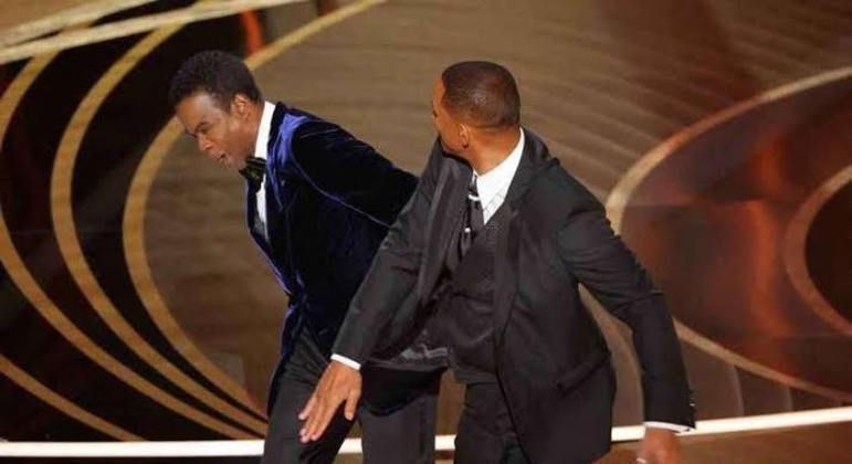 O ator Chris Rock depois de acabar de dar um tapa no comediante Chris Rock, apresentador do Oscar 2022. Os dois estão de smoking, o de Rock com paletó de veludo azul e calça preta, o de Smith com calça e paletó pretos.