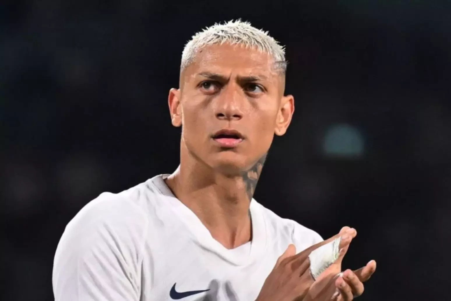 Richarlison, jogador de futebol, com cabelo loiro platinado e tatuagens no pescoço, veste camisa branca da Nike e aplaude com as mãos enfaixadas, olhando para cima com expressão séria em um fundo escuro