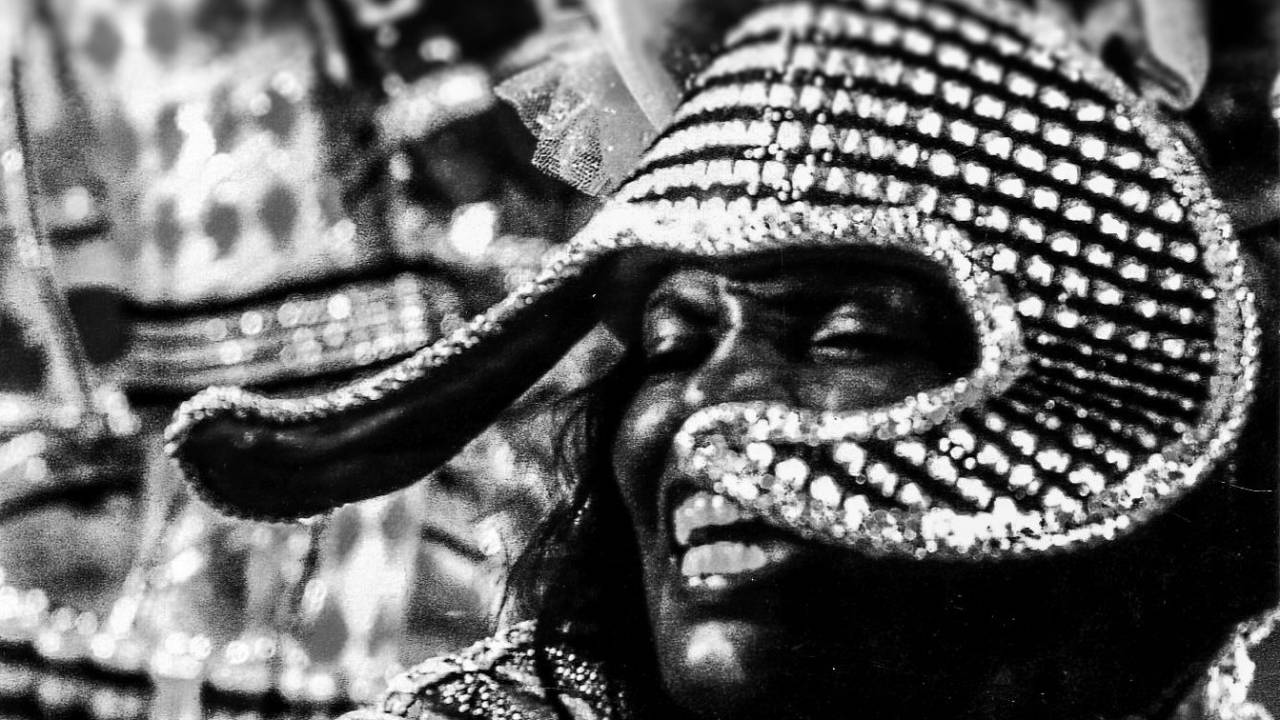 Integrante de escola de samba, mulher negra, com adereço na cabeça, sorrindo. em foto em preto e branco.