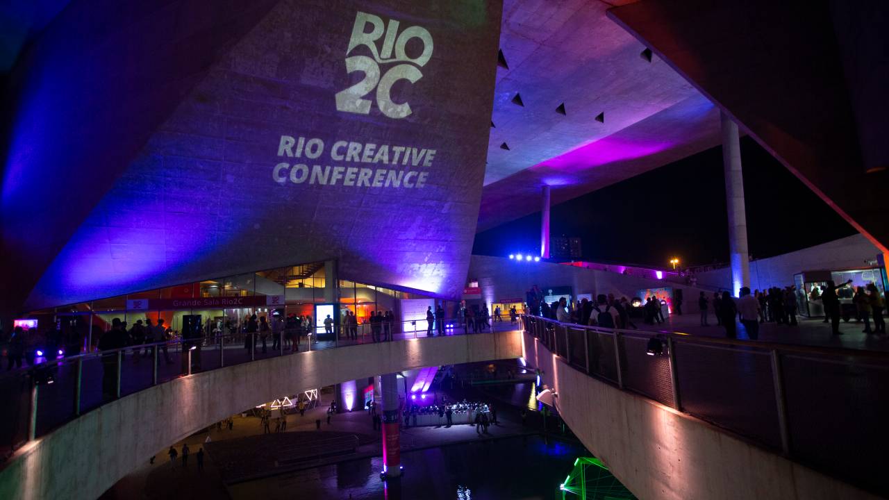 Foto de cima da Cidade das Artes com iluminação roxa e o nome Rio2C - Rio Creative Conference projetado em branco em uma parte do concreto do lugar.