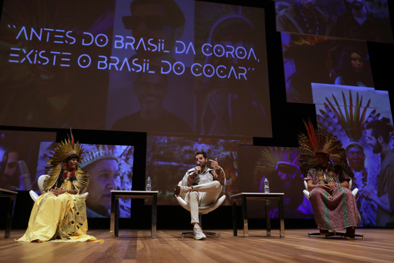 Celia Xakriabá, Alok e cacique Mapu Huni Kuî sentados em um auditório durante um painel no Rio2C. Atrás deles, está projetada a frase: Antes do Brasil da coroa, existe o Brasil do cocar, nome do painel