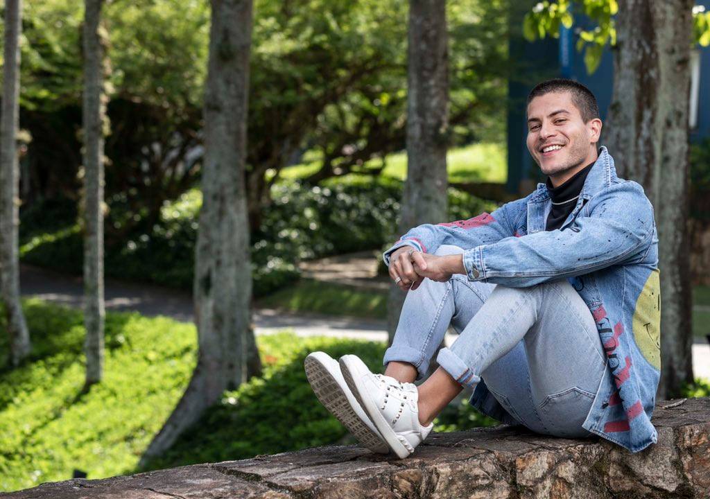 Arthur Aguiar de calça e jaqueta jeans, usando tênis branco sem meia, sentado em uma mureta de pedra no Projac, cercado de verde.