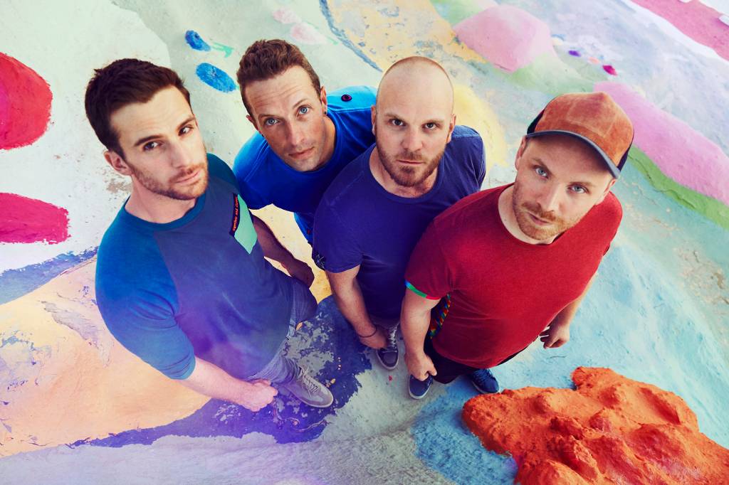 Os quatro integrantes do Coldplay estão olhando para cima, sobre um chão colorido. Os três da esquerda usam camisetas azuis, e o da direita usa camiseta vermelha e boné.