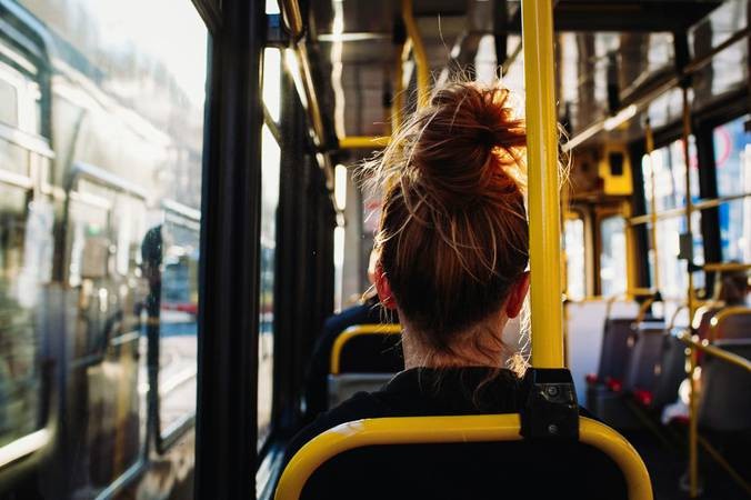 Pessoa com cabelo castanho avermelhado preso em coque, vista de costas, sentada em ônibus com assento preto e barra amarela. A luz do sol entra pela janela lateral, refletindo no vidro e iluminando o cabelo