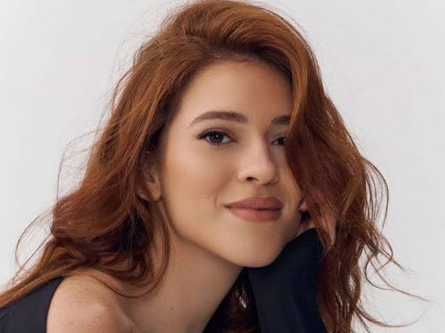 Mulher jovem de cabelos ruivos ondulados, sorrindo levemente, com a mão direita apoiada no queixo, vestindo blusa preta, em fundo branco