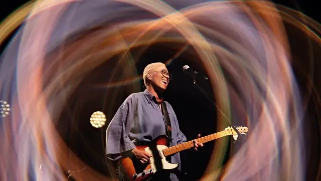 Pessoa de cabelo curto e loiro, óculos redondos, camisa azul folgada, sorrindo e tocando guitarra elétrica em palco escuro com luzes circulares coloridas ao fundo