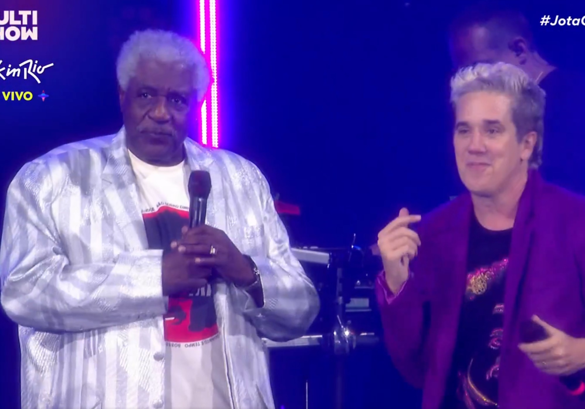 Dois homens em um palco iluminado por luzes azuis e roxas. À esquerda, um homem negro de cabelos grisalhos, blazer prateado listrado e camiseta branca com estampa preta e vermelha, segura um microfone. À direita, um homem branco de cabelo loiro platinado e blazer roxo, sorri e faz um gesto com a mão direita