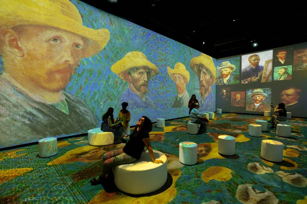 Sala com pessoas sentadas em pufes e projeções com telas de Van Gogh em duas paredes e no chão.
