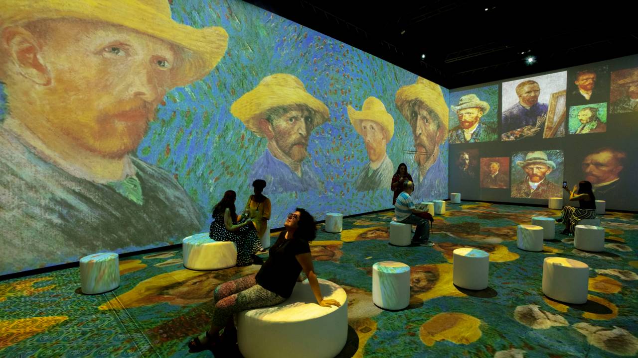 Sala com pessoas sentadas em pufes e projeções com telas de Van Gogh em duas paredes e no chão.