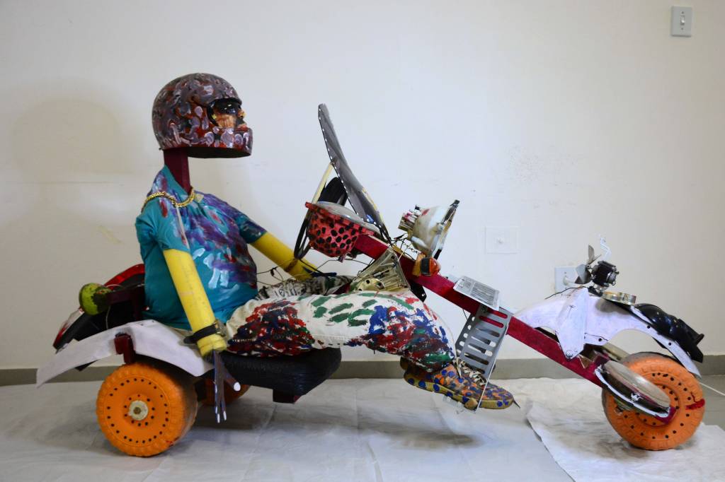 Escultura traz figura humana de capacete sentada em um carrinho que lembra um kart.