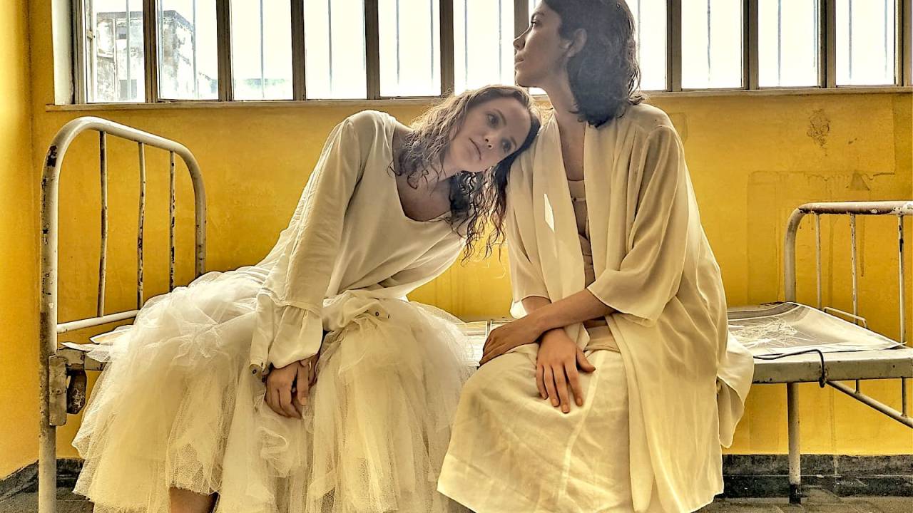 As atrizes Danielle Oliveira e Maria Augusta Montera vestidas de roupas brancas em cima de uma cama.