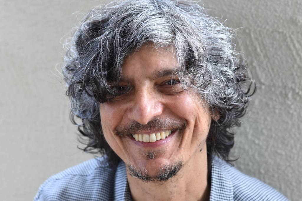 O cantor e compositor Pedro Luís está olhando para a câmera e sorrindo. Ele usa camisa cinza quadriculada.