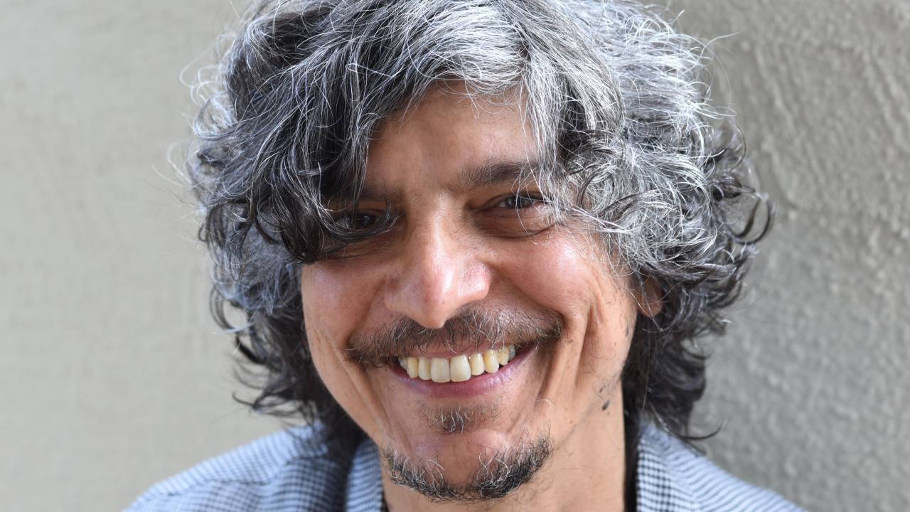 O cantor e compositor Pedro Luís está olhando para a câmera e sorrindo. Ele usa camisa cinza quadriculada.