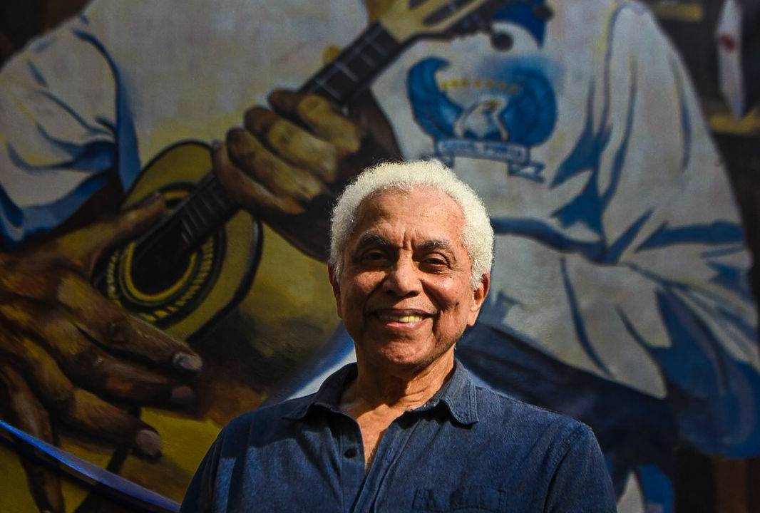 Foto mostra o cantor Paulinho da Viola em frente a muro pintado com seu retrato
