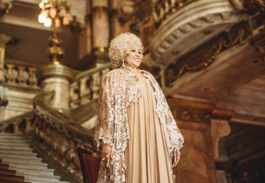 A cantora Alcione veste vestido bege lindo com casaco comprido de renda bege. Ela está nas escadarias do Theatro Municipal.