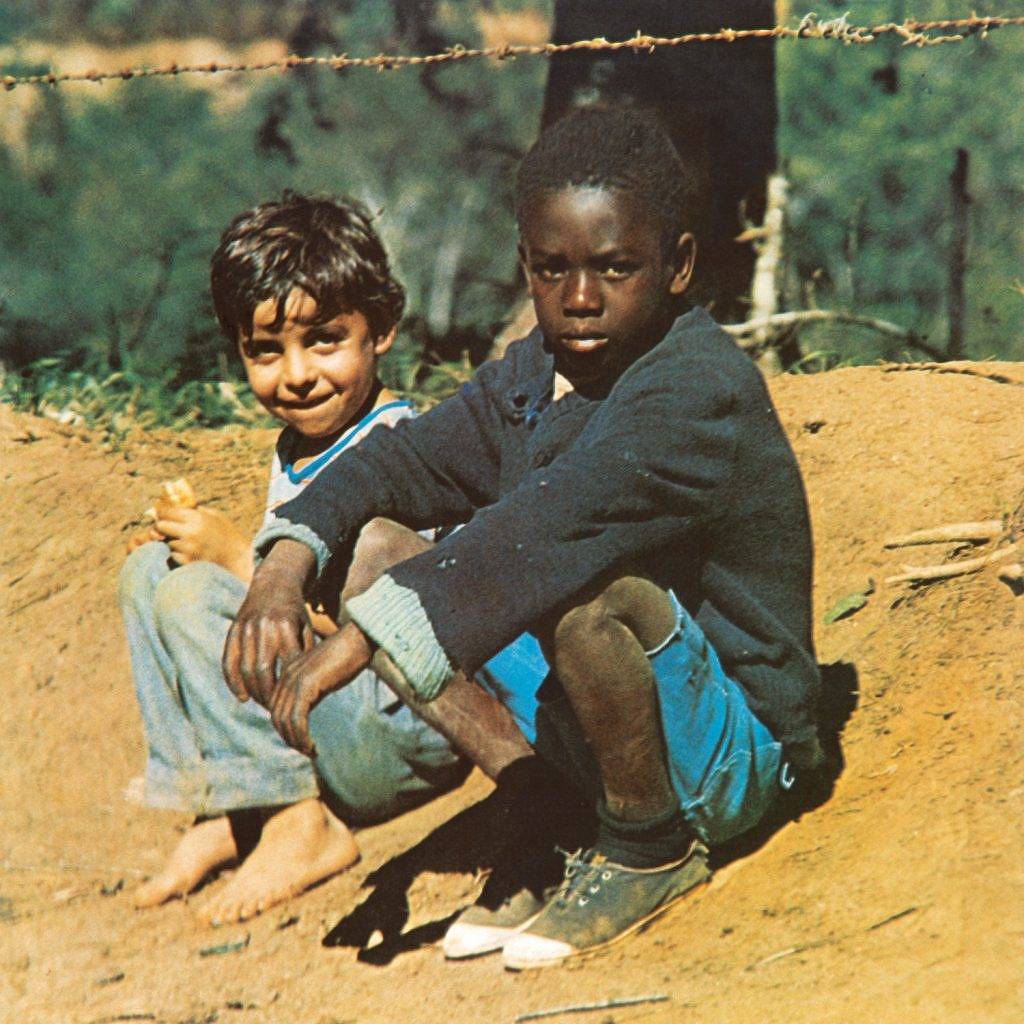 Dois meninos estão sentados em um barranco. À esquerda, Tonho, um menino branco, de cabelos escuros e lisos, de calça jeans. À direita, na frente, Cacau, um menino negro, de cabelo curto, usando bermuda azul-turquesa e blusa de manga comprida azul-escuro.