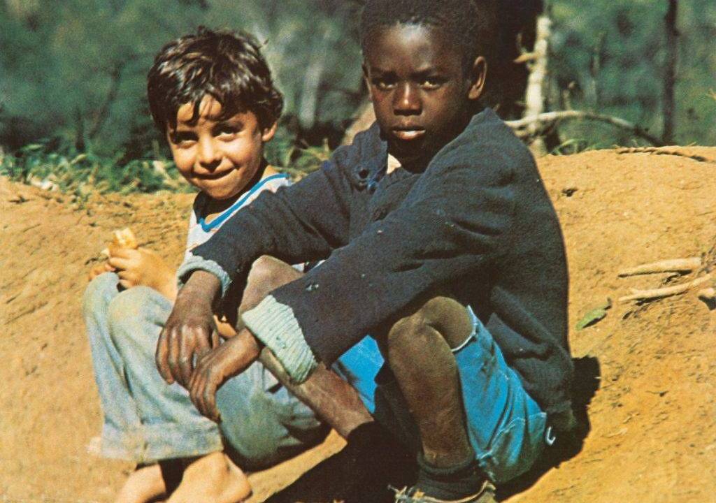 Dois meninos estão sentados em um barranco. À esquerda, Tonho, um menino branco, de cabelos escuros e lisos, de calça jeans. À direita, na frente, Cacau, um menino negro, de cabelo curto, usando bermuda azul-turquesa e blusa de manga comprida azul-escuro.