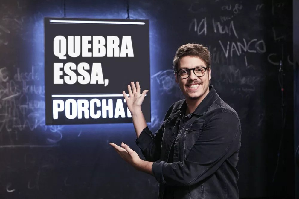 Foto mostra o ator Fábio Porchat apontando para uma placa de luz branca neon com fundo preto escrita "Quebra essa, Porchat?"