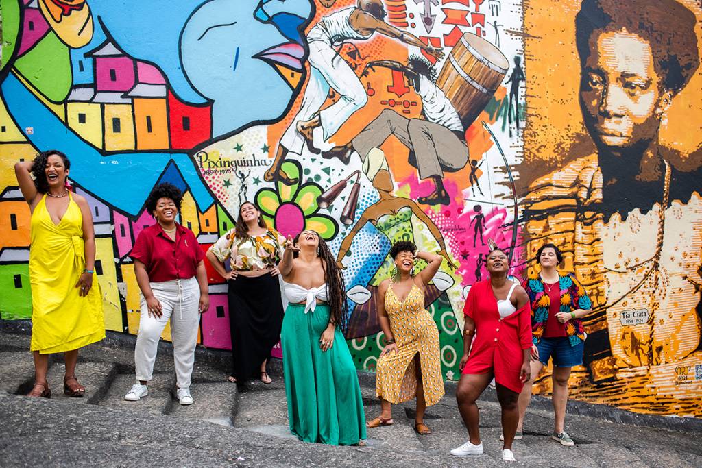 A sete integrantes da roda de samba da Moça Prosa de pé, sorrindo, com uma parede grafitada ao fundo.