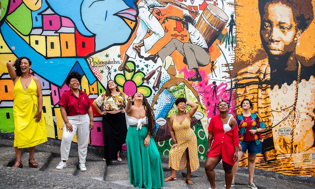 A sete integrantes da roda de samba da Moça Prosa de pé, sorrindo, com uma parede grafitada ao fundo.