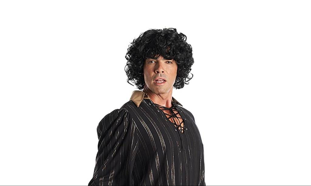 O ator Márcio Louzada caracterizado como Sidney Magal, com peruca de cabelos pretos cacheados, camisa preta com amarração na frente e gola bege.