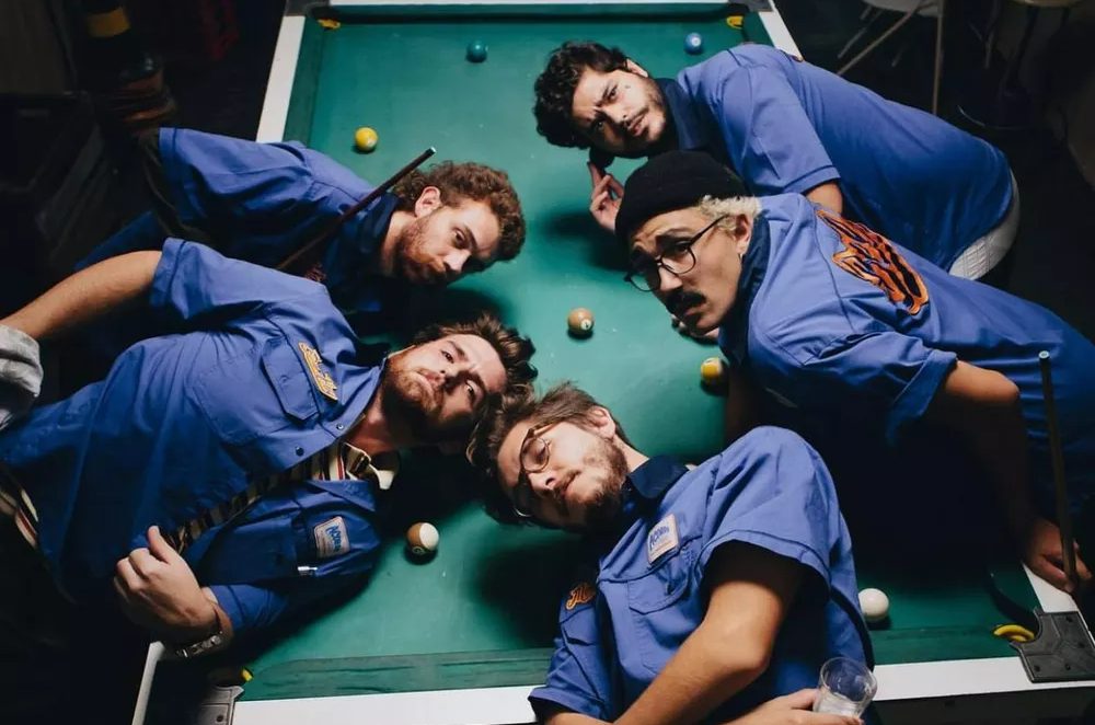 Os cinco integrantes do grupo jovem Dionisio usando camisa azul de manga curta e deitados em uma mesa de sinuca.