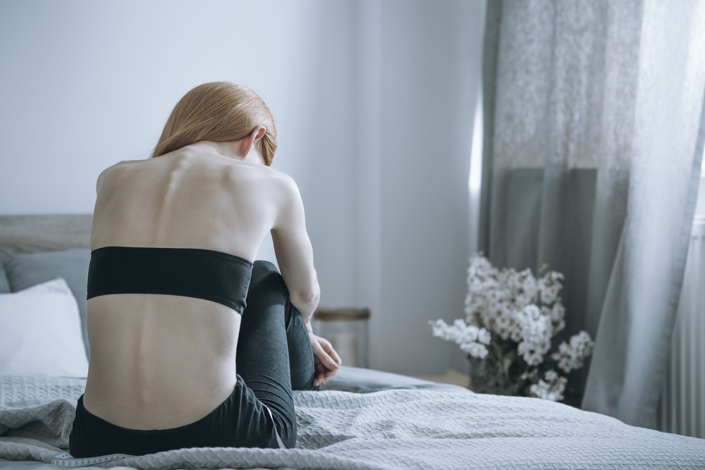 Jovem de magreza acentuada sentada de costas na cama.