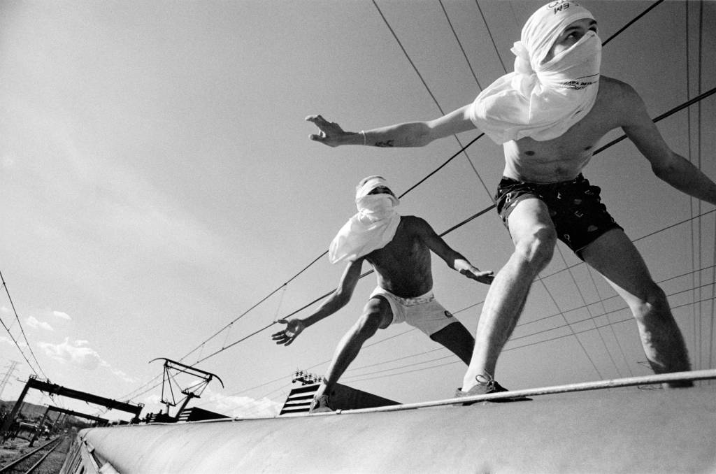 Foto em preto e branco de dois jovens com a cabeça cobertura com camisetas "surfando" um trem -