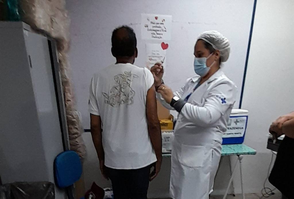 O cantor e compositor Jorge Ben Jor está de costas, vestindo calça jeans e blusa branca com estampa de São Jorge, enquanto é vacinado pela enfermeira do posto de saúde, que está de lado, vestida de branco, usando touca e máscara.