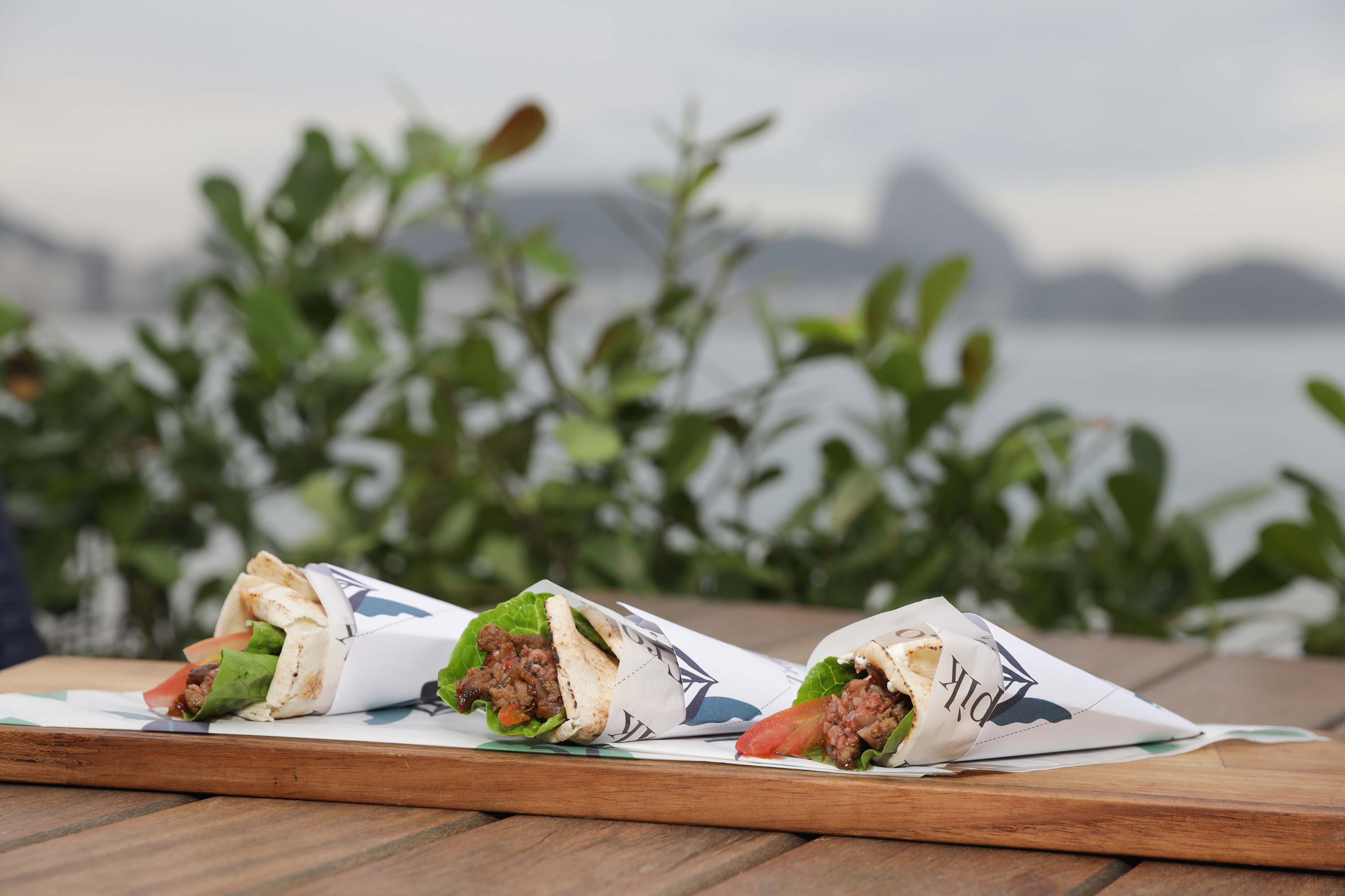 Foto mostra kebabs em cima de uma mesa. Ao fundo da foto está a praia de copacabana