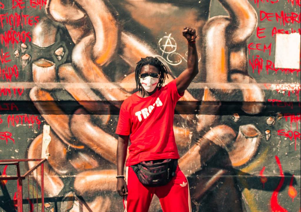 Schackal é um homem negro de tranças curtas. Está de calça Adidas e camiseta com a palavra Tropa escrita, ambas vermelhas. Está de máscara branca e com o punho erguido. Ao fundo, um grafite com correntes.