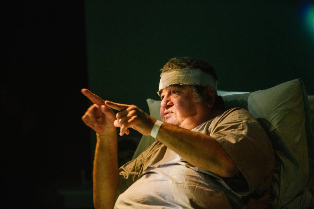 O ator Otávio Augusto está apoiado em uma cama, gesticulando, e tem uma atadura em torno da testa.